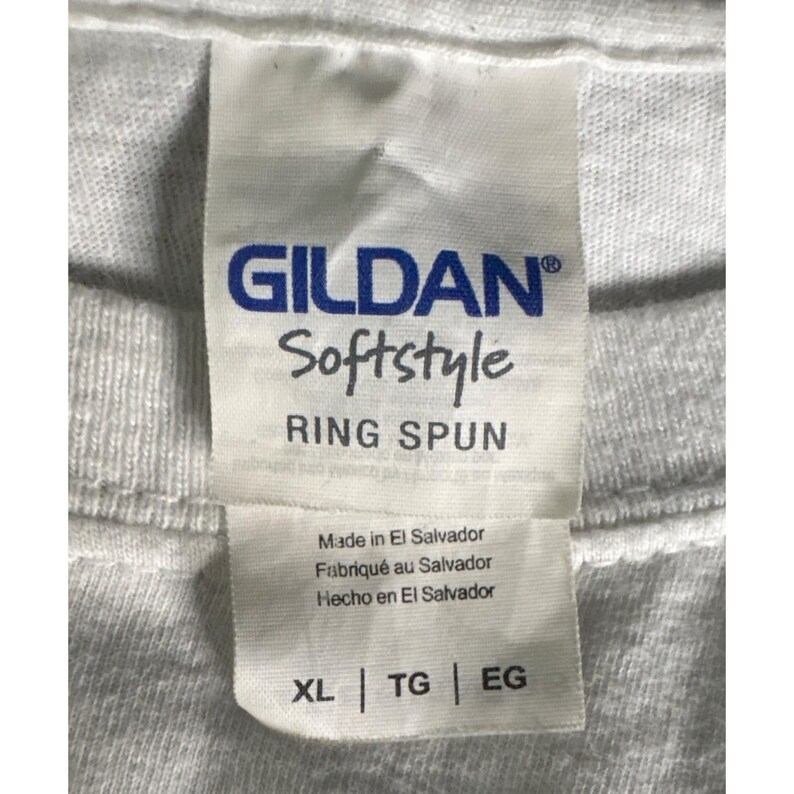 May include: Close-up of a white Gildan Softstyle t-shirt tag. The tag includes the brand name, fabric type, and country of origin. The tag also shows the size XL, TG, and EG.