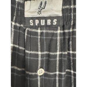 May include: Black and white checked pajama trousers with a button fastening. The waistband features a tag with the word "SPURS" and a team logo. The trousers have a classic checked pattern.