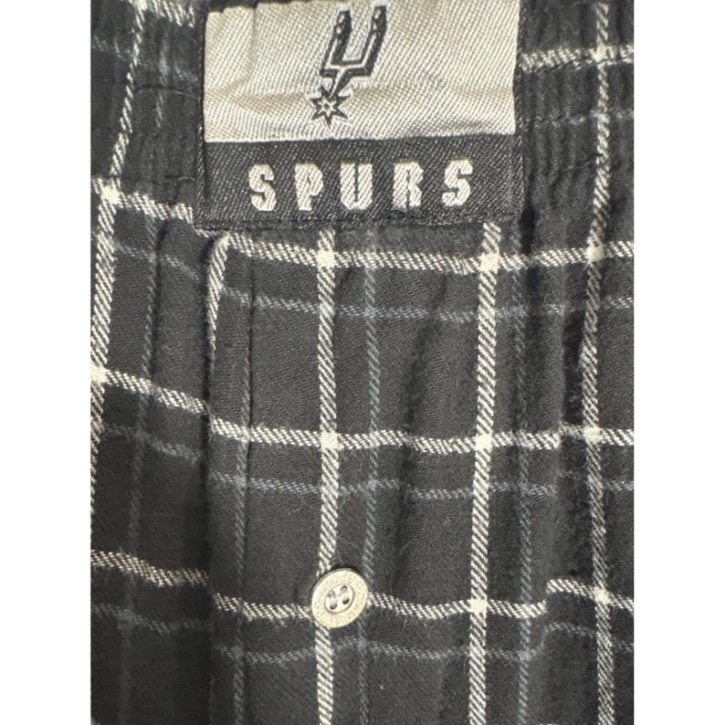 May include: Black and white checked pajama trousers with a button fastening. The waistband features a tag with the word "SPURS" and a team logo. The trousers have a classic checked pattern.