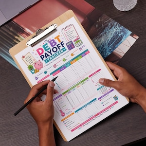 May include: A wooden clipboard holding a printed My Debt Payoff Tracker sheet with colorful sections, a pink piggy bank illustration, a calculator icon, and various financial tracking tables, being written on with a black pen.