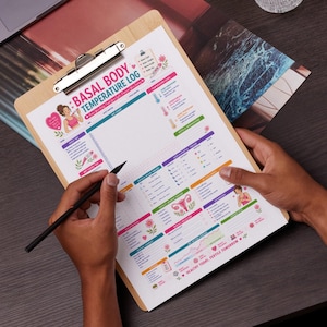 Op de afbeelding: Een houten clipboard met een afgedrukte "Basal Body Temperature Log" grafiek. De grafiek bevat secties voor het volgen van temperatuur, cervixslijm en andere vruchtbaarheidsindicatoren. Een zwarte pen wordt boven de grafiek gehouden.