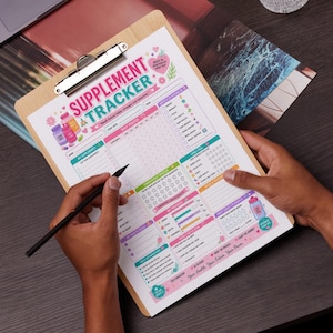May include: A wooden clipboard with a colorful "Supplement Tracker" sheet. The sheet includes sections for supplements, daily tracking, goals, and affirmations. A black pen is held over the sheet, ready to record information. The design is bright and cheerful.