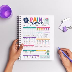 Puede incluir: Un cuaderno con espiral titulado "Pain Tracker" con gráficos coloridos. El cuaderno incluye secciones para rastrear los niveles de dolor, los posibles desencadenantes y los conocimientos diarios. Un bolígrafo morado se sostiene cerca. Medidas en cm.