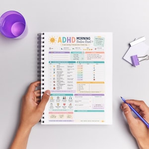 May include: A spiral-bound planner with the text "ADHD Morning Routine Reset" in colourful lettering. The planner's pages are filled with checklists, charts, and icons for daily planning and goal setting. A purple cup and pen are also visible.