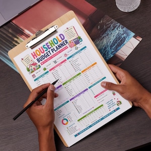 May include: A wooden clipboard holding a printed household budget planner sheet with colorful sections, icons, and text including Household Budget Planner, Income Tracker, Variable Expenses, and Savings Goals, with a black pen resting on the paper.