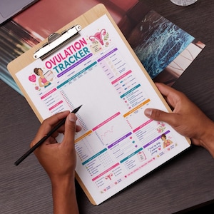 May include: A wooden clipboard displaying an ovulation tracker chart with colourful graphics and the text "Ovulation Tracker." A person holds the clipboard and a black pen, ready to write. The chart includes sections for tracking cycle data. Metric measurements.