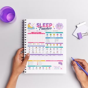 May include: A spiral-bound sleep tracker journal with the title "SLEEP TRACKER" in pink and purple. The journal features sections for sleep calendar, sleep quality, and daily sleep log. A purple pen is held in the hand.