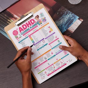 May include: A wooden clipboard with an ADHD Cleaning Planner. The planner has colorful sections for daily, weekly, and monthly cleaning tasks, with a focus on cleaning by zone and tracking progress. The text on the planner reads "ADHD Cleaning Planner: Focus, Reset, Clean, Feel Good."