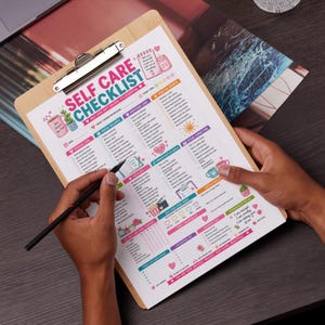 Peut inclure: Un presse-papiers en bois avec une "Self Care Checklist" colorée, divisée en catégories et cases à cocher. La liste comprend des sections pour le bien-être physique, émotionnel et social. Un stylo noir est tenu au-dessus de la liste.