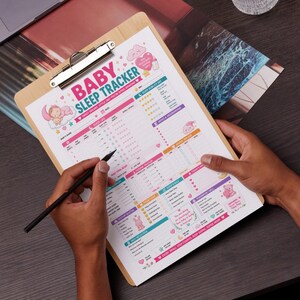 Könnte beinhalten: Ein Holz-Klemmbrett mit einem "Baby Sleep Tracker"-Blatt mit rosa, türkis und gelben Akzenten. Das Blatt enthält Abschnitte für Schlaflogbücher, Fütterung und tägliche Reflexionen. Eine Hand hält einen schwarzen Stift und markiert das Blatt.