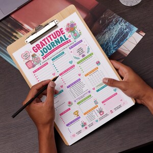 May include: A wooden clipboard holds a colorful gratitude journal with the title "Gratitude Journal" in pink and white. The journal features sections for daily reflections, with floral and heart illustrations. A hand holds a pencil, ready to write.