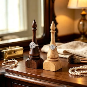 May include: Two wooden ring holders, one dark brown and one light tan, each featuring a pointed top and a square base. Each holder displays a silver ring with a clear, oval-shaped stone set in the center. The holders are made of wood.