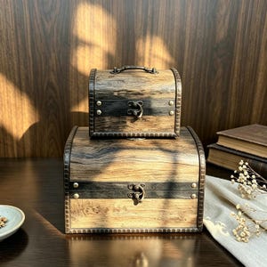 Puede incluir: Dos cofres de almacenamiento de madera apilados con tapa curva. Cada cofre presenta un acabado de veta de madera marrón claro, bandas de metal oscuro, tachuelas decorativas y un cierre central de metal. El cofre pequeño incluye un asa.