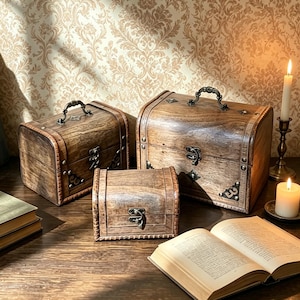 May include: Three wooden storage chests with arched lids, dark brown wood grain, and antique-style metal hardware. Each chest features a front latch, decorative corner accents, and a top handle on the two larger boxes, arranged in a group.