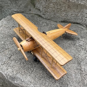 Pode incluir: Um modelo de biplano de madeira com acabamento castanho claro e um padrão de grelha nas asas. A aeronave inclui uma hélice montada na frente, dois conjuntos de asas sobrepostas ligadas por suportes verticais, cabos de arame preto e cauda.