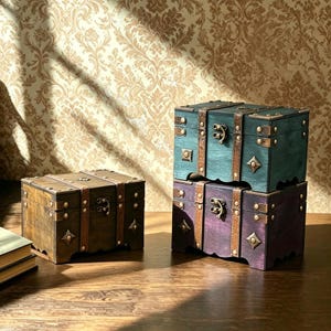 May include: Three wooden storage chests with metal latches and decorative corner accents. One chest is dark brown, one is teal, and one is purple. Each chest features vertical brown straps, metal studs, and a hinged lid with a front locking mechanism.