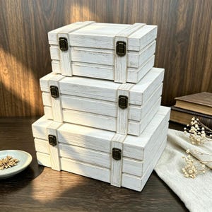 Könnte beinhalten: Ein Stapel aus drei rechteckigen Aufbewahrungsboxen aus Holz mit weißem Used-Look-Finish. Jede Box verfügt über zwei vertikale Zierleisten und zwei bronzefarbene Metallverschlüsse an der Vorderseite. Die Boxen sind in einer Stufenform angeordnet.