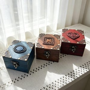 Könnte beinhalten: Drei quadratische Holzkisten mit Metallverschlüssen und Eckverzierungen. Die blaue Kiste hat ein Muschelemblem, die braune Kiste eine quadratische Schmetterlingsschnitzerei und die rote Kiste zeigt eine herzförmige Schnitzerei mit einem Schmetterling.