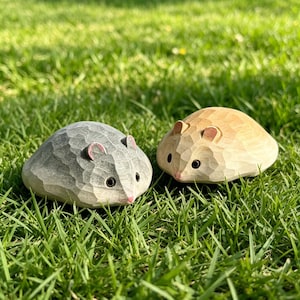 Könnte beinhalten: Zwei kleine, handgeschnitzte Holzfiguren in Form von abgerundeten Nagetieren. Eine Figur ist in einem gedeckten Grauton gehalten, während die andere eine natürliche, hellbraune Holzmaserung aufweist. Beide haben geschnitzte Ohren, dunkle Augen.