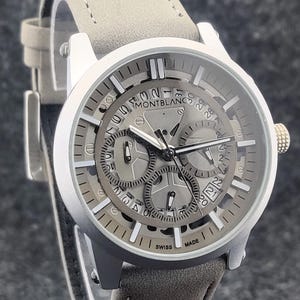 May include: A silver Montblanc watch with a grey face and a grey leather strap. The watch face has multiple subdials and the text "MONTBLANC" at the top. The watch is marked "SWISS MADE".