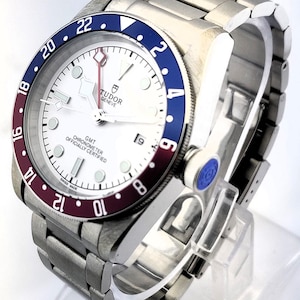 May include: A stainless steel wristwatch with a white face, a blue and burgundy bezel, and a date display at the 3 o'clock position. The watch has a metal band and the text "TUDOR GENEVE GMT CHRONOMETER OFFICIALLY CERTIFIED SWISS MADE" is visible.