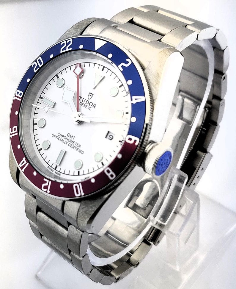 May include: A stainless steel wristwatch with a white face, a blue and burgundy bezel, and a date display at the 3 o'clock position. The watch has a metal band and the text "TUDOR GENEVE GMT CHRONOMETER OFFICIALLY CERTIFIED SWISS MADE" is visible.
