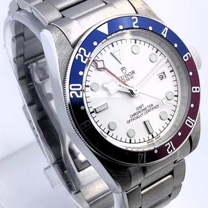 May include: A stainless steel wristwatch with a white dial, a blue and burgundy bezel, and a matching stainless steel band. The watch face features the brand name "TUDOR GENEVE" and the text "GMT CHRONOMETER OFFICIALLY CERTIFIED SWISS MADE".