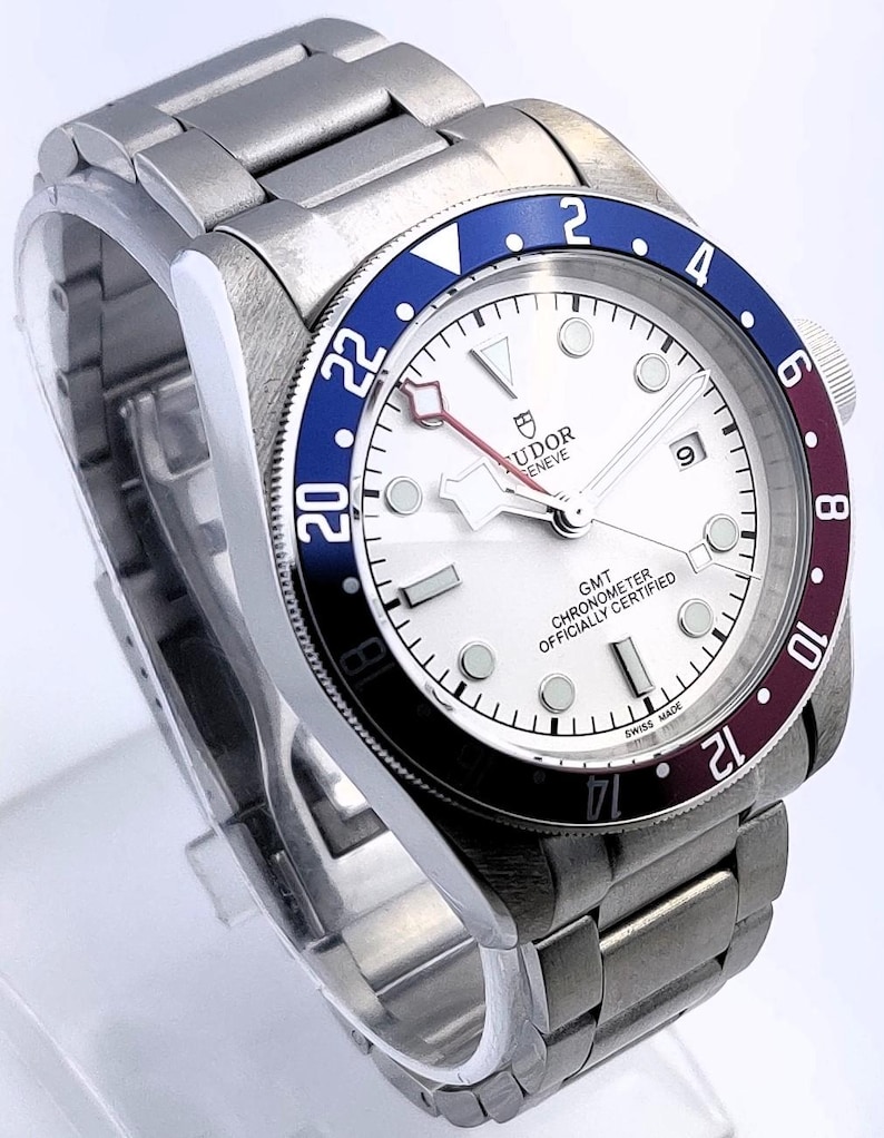 May include: A stainless steel wristwatch with a white dial, a blue and burgundy bezel, and a matching stainless steel band. The watch face features the brand name "TUDOR GENEVE" and the text "GMT CHRONOMETER OFFICIALLY CERTIFIED SWISS MADE".