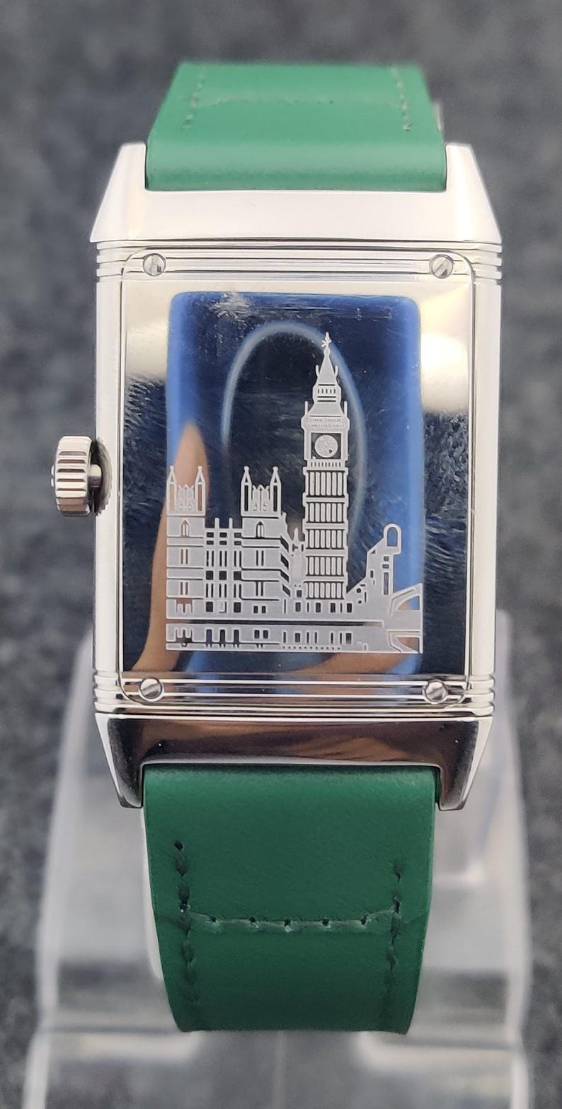 May include: A rectangular silver wristwatch with a green leather band. The watch face features an etched image of the Big Ben clock tower and other buildings. The watch has a silver crown and a green leather strap.