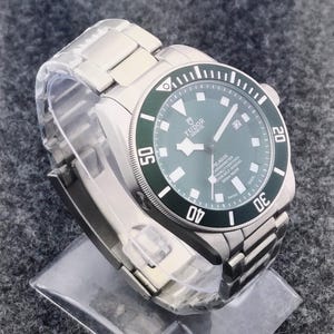May include: A stainless steel wristwatch with a green bezel and a matching green dial. The watch features a date display at the 3 o'clock position and a stainless steel bracelet. The watch has the text "TUDOR GENEVE PELAGOS" on the dial.