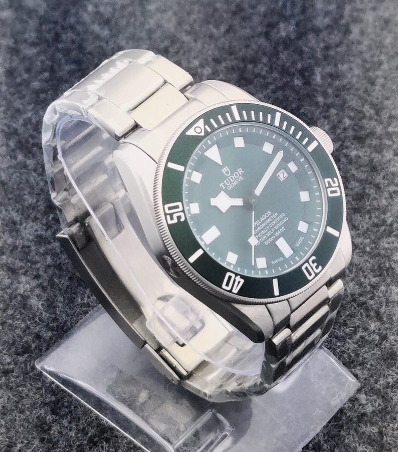 May include: A stainless steel wristwatch with a green bezel and a matching green dial. The watch features a date display at the 3 o'clock position and a stainless steel bracelet. The watch has the text "TUDOR GENEVE PELAGOS" on the dial.