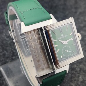 May include: A rectangular wristwatch with a green face and a green leather band. The watch has silver-colored accents and a small seconds dial. The band is stitched with white thread. The watch face has the text "Jaeger-LeCoultre".