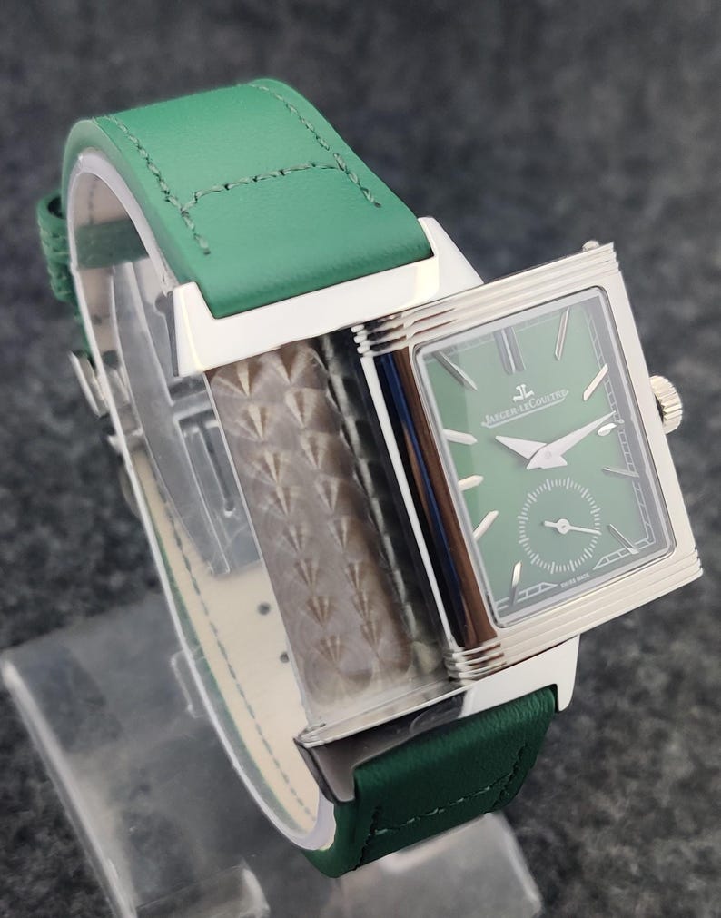 May include: A rectangular wristwatch with a green face and a green leather band. The watch has silver-colored accents and a small seconds dial. The band is stitched with white thread. The watch face has the text "Jaeger-LeCoultre".