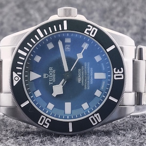 May include: A stainless steel wristwatch with a blue dial and a black rotating bezel. The watch features white markers and hands, with the brand name "TUDOR GENEVE" visible on the dial. The watch band is also stainless steel.