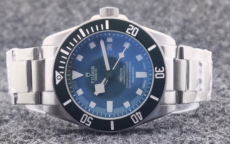May include: A stainless steel wristwatch with a blue dial and a black rotating bezel. The watch features white markers and hands, with the brand name "TUDOR GENEVE" visible on the dial. The watch band is also stainless steel.