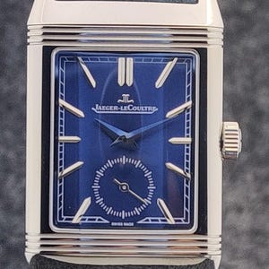 May include: A rectangular wristwatch with a blue face and a blue leather band. The watch face features gold-colored hands and markers, with the brand name "JAEGER-LECOULTRE" displayed prominently. The watch has a silver-colored case and a small seconds dial.