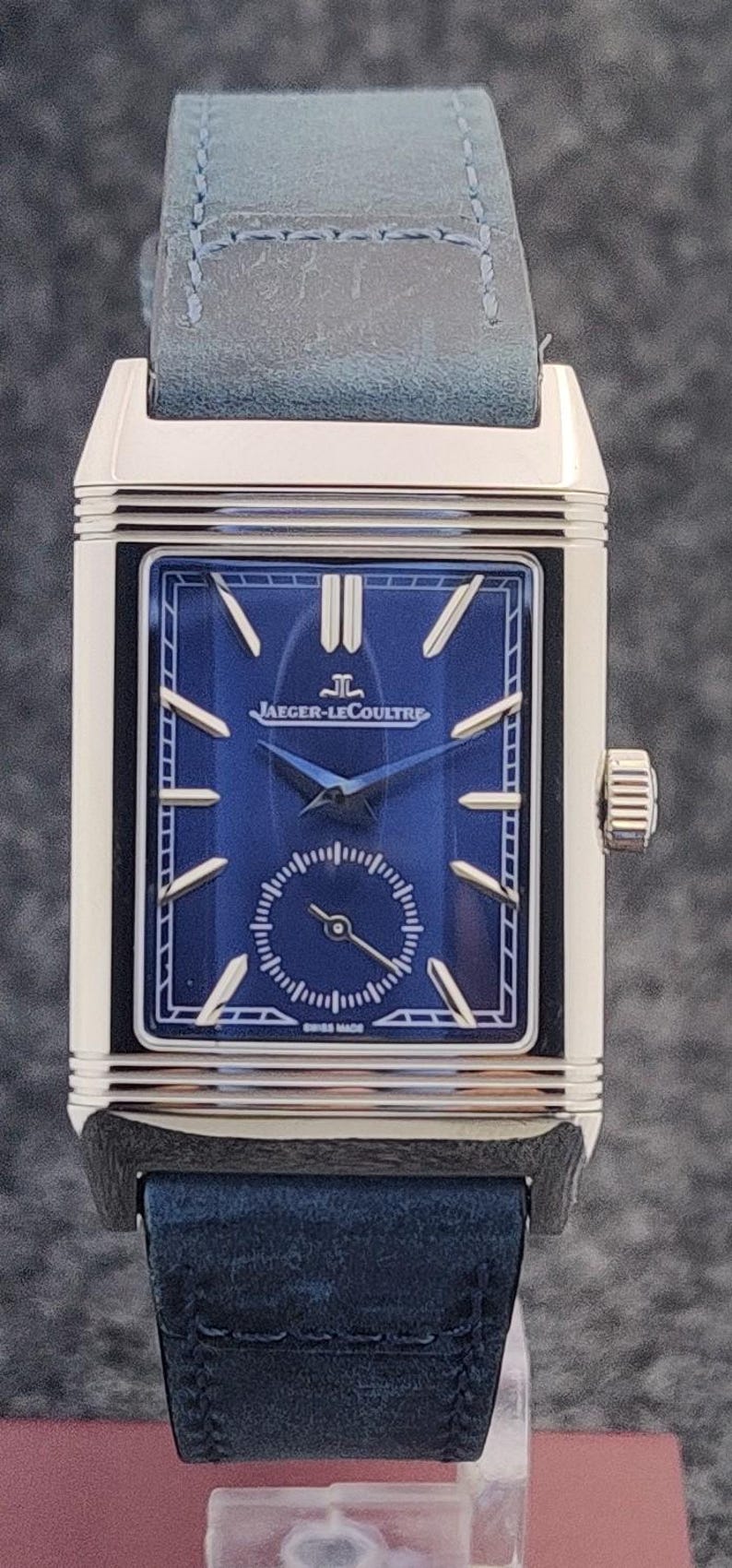 May include: A rectangular wristwatch with a blue face and a blue leather band. The watch face features gold-colored hands and markers, with the brand name "JAEGER-LECOULTRE" displayed prominently. The watch has a silver-colored case and a small seconds dial.
