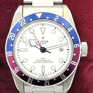 Puede incluir: Un reloj de pulsera de acero inoxidable con esfera blanca, bisel azul y burdeos, y correa plateada. La esfera del reloj presenta el texto "TUDOR GENEVE GMT CHRONOMETER OFFICIALLY CERTIFIED SWISS MADE". El reloj se muestra sobre una superficie roja.