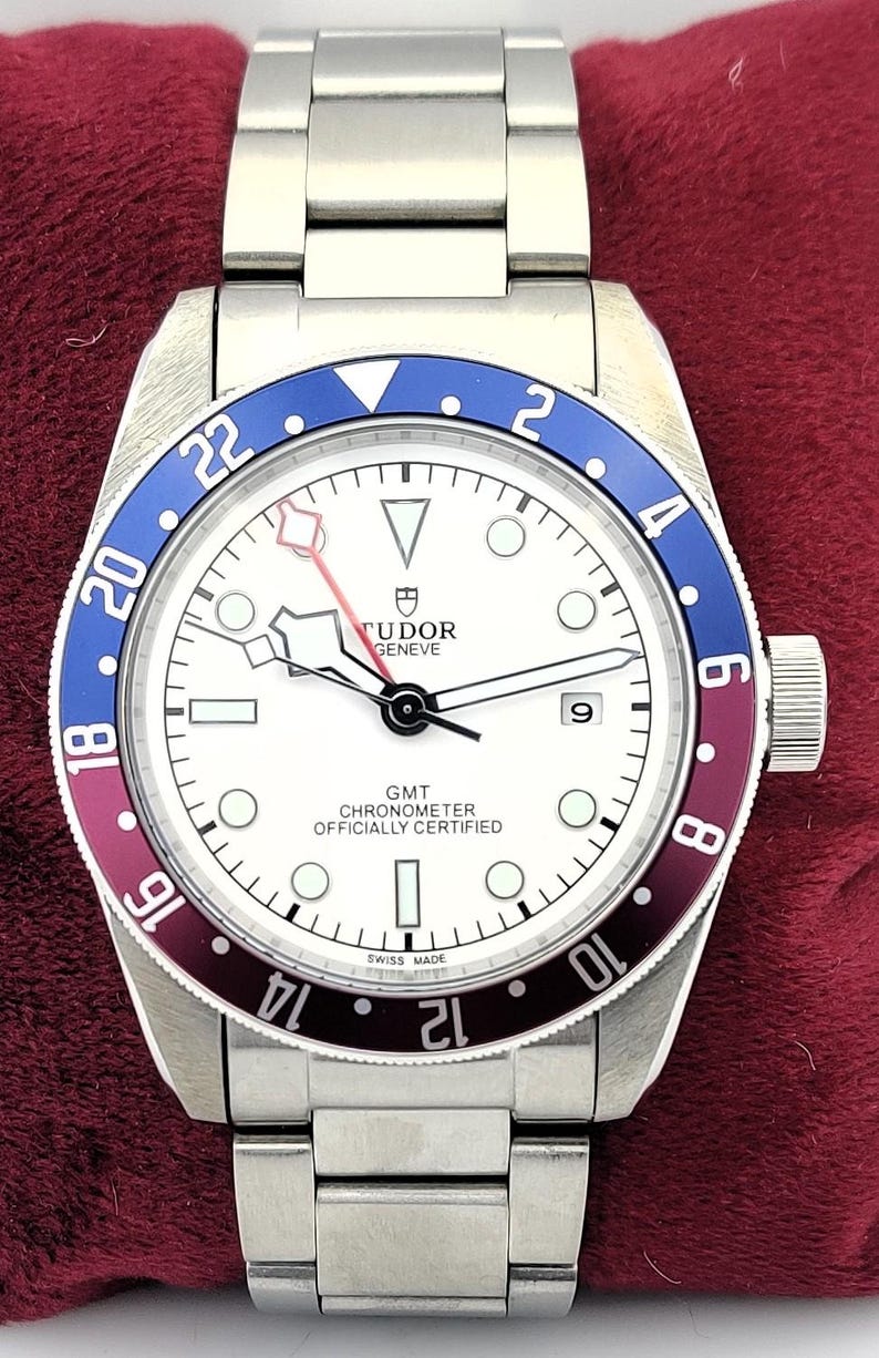 May include: A stainless steel wristwatch with a white face, a blue and burgundy bezel, and a silver band. The watch face features the text "TUDOR GENEVE GMT CHRONOMETER OFFICIALLY CERTIFIED SWISS MADE". The watch is displayed on a red surface.