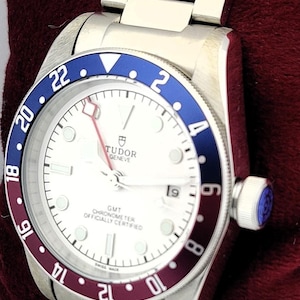May include: A silver-toned wristwatch with a white face and a blue and burgundy bezel. The watch features a stainless steel band and a crown with a blue accent. The watch face displays the brand name "TUDOR GENEVE".