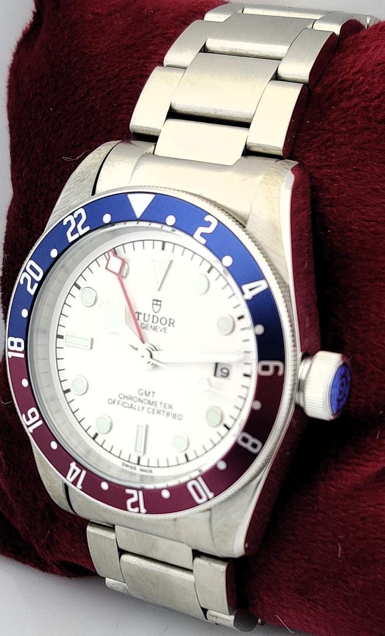 May include: A silver-toned wristwatch with a white face and a blue and burgundy bezel. The watch features a stainless steel band and a crown with a blue accent. The watch face displays the brand name "TUDOR GENEVE".
