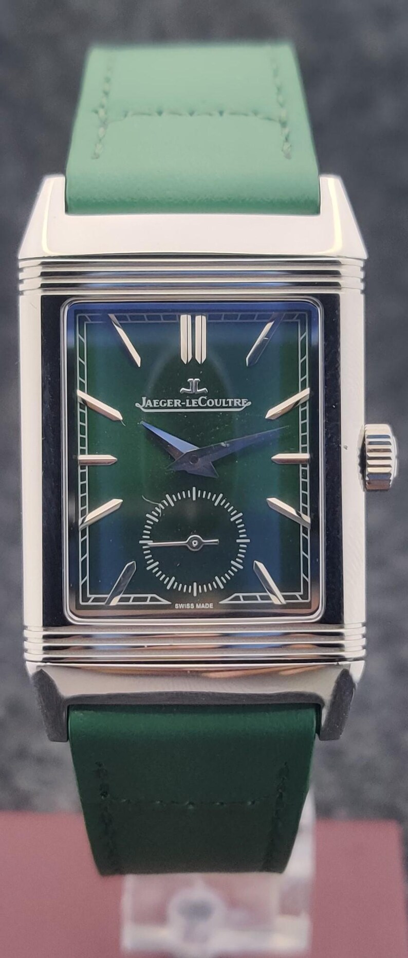 May include: A rectangular Jaeger-LeCoultre watch with a green dial and a green leather strap. The watch face has silver hands and markers, with a small seconds dial at the bottom. The watch is Swiss made.