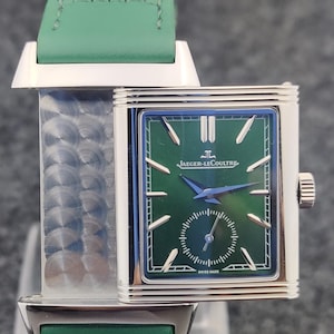 May include: A rectangular wristwatch with a green face and a green leather strap. The watch face features the Jaeger-LeCoultre logo and a small seconds dial. The watch has a silver-colored case and a green leather strap.