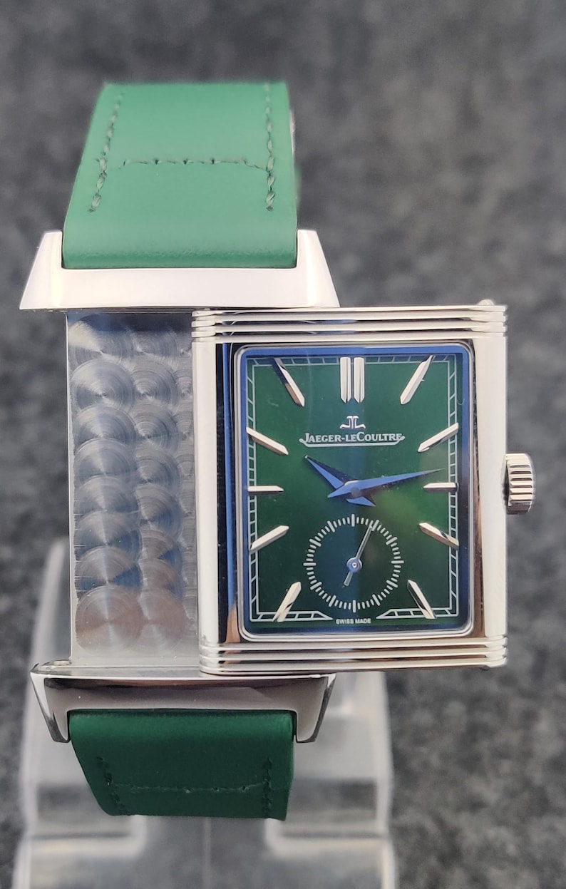 May include: A rectangular wristwatch with a green face and a green leather strap. The watch face features the Jaeger-LeCoultre logo and a small seconds dial. The watch has a silver-colored case and a green leather strap.