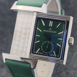 May include: A rectangular wristwatch with a green face and a green leather band. The watch has silver-colored accents and the brand name "Jaeger-LeCoultre" is visible on the watch face. The watch band is approximately 0.75 inches wide.