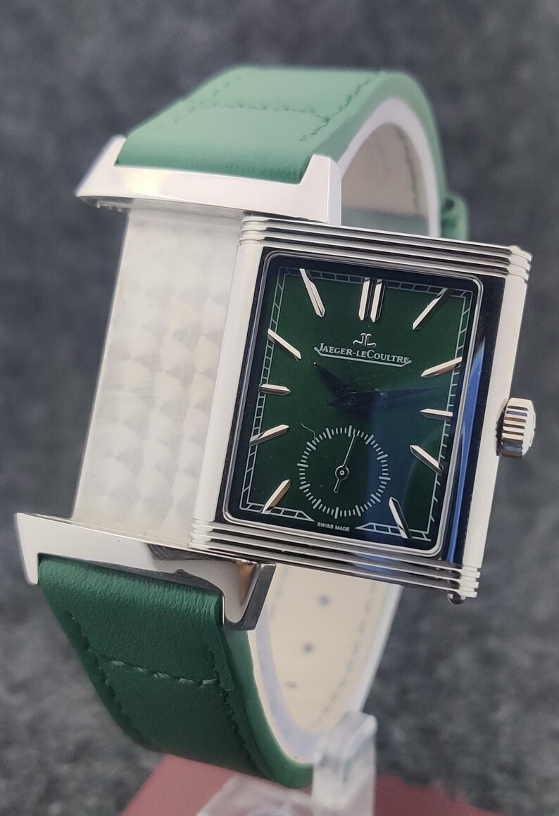 May include: A rectangular wristwatch with a green face and a green leather band. The watch has silver-colored accents and the brand name "Jaeger-LeCoultre" is visible on the watch face. The watch band is approximately 0.75 inches wide.