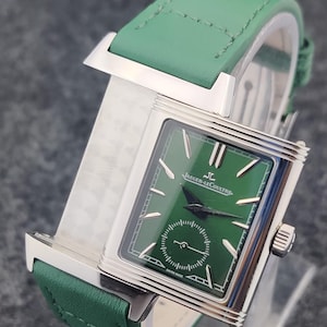 May include: A luxury wristwatch with a green leather band and a rectangular silver case. The watch face is green with silver hands and markers, and the brand name "Jaeger-LeCoultre" is displayed. The watch has a small seconds dial.
