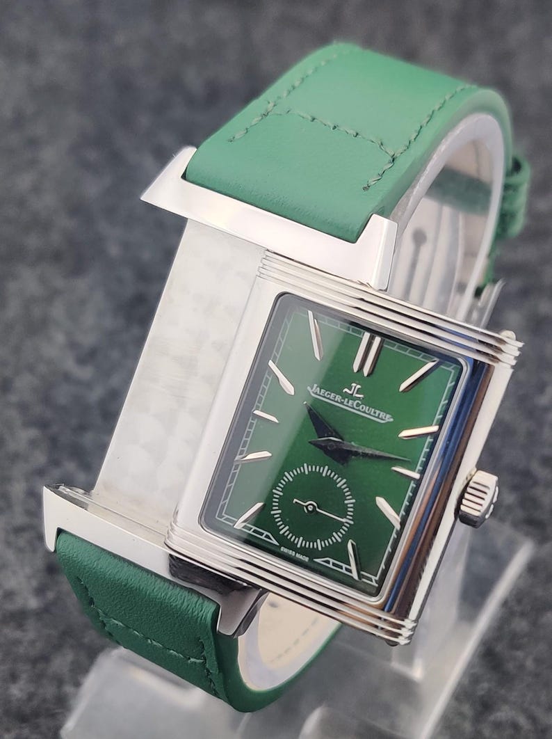 May include: A luxury wristwatch with a green leather band and a rectangular silver case. The watch face is green with silver hands and markers, and the brand name "Jaeger-LeCoultre" is displayed. The watch has a small seconds dial.