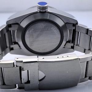 May include: A stainless steel wristwatch with a silver band and a blue crown. The watch face is not visible, but the back of the watch shows the brand name and the words "GENEVE" and "TUDOR". The watch band is closed.