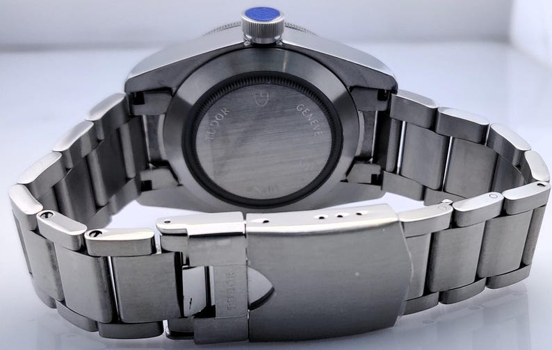 May include: A stainless steel wristwatch with a silver band and a blue crown. The watch face is not visible, but the back of the watch shows the brand name and the words "GENEVE" and "TUDOR". The watch band is closed.
